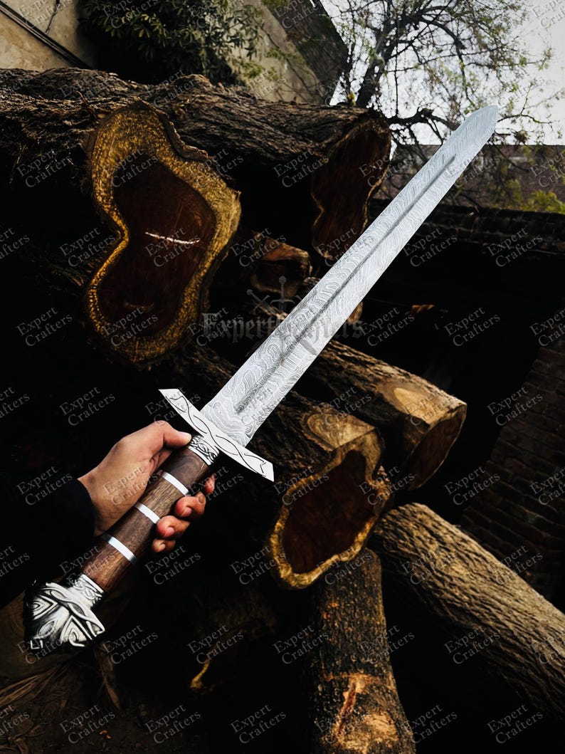 Op de afbeelding: Een lang zwaard met een patroon van staal en een houten handvat. De handgreep heeft zilveren accenten en een decoratieve pommel. Het zwaard wordt getoond tegen een achtergrond van grote, gezaagde boomstammen.