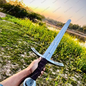 May include: A silver-colored sword with a cross-shaped hilt and a dark brown handle. The blade has the word "VINCIT" on one side and "CHRISTUS" on the other. The sword is held against a backdrop of green grass and a sunset.