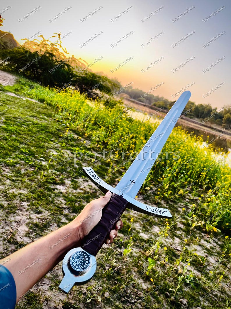 May include: A silver-colored sword with a cross-shaped hilt and a dark brown handle. The blade has the word "VINCIT" on one side and "CHRISTUS" on the other. The sword is held against a backdrop of green grass and a sunset.