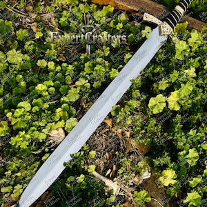 May include: A long, silver sword with a decorative gold hilt and a black handle, resting on a bed of green foliage. The sword has a red gem at the pommel. The text "Expert Crafters" is visible on the blade.