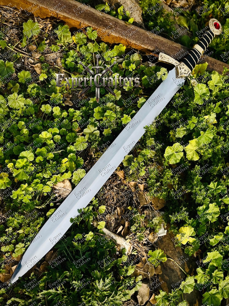 May include: A long, silver sword with a decorative gold hilt and a black handle, resting on a bed of green foliage. The sword has a red gem at the pommel. The text "Expert Crafters" is visible on the blade.