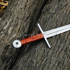 May include: A medieval sword with a silver blade and crossguard, a brown leather-wrapped handle, and a silver pommel featuring a black and white emblem. The sword rests on a textured, brown wooden surface.