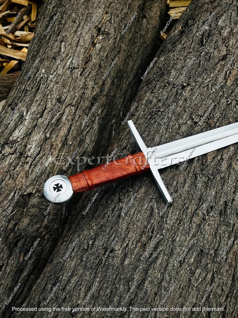 May include: A medieval sword with a silver blade and crossguard, a brown leather-wrapped handle, and a silver pommel featuring a black and white emblem. The sword rests on a textured, brown wooden surface.