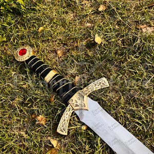 May include: A decorative sword with a silver blade and gold-colored hilt. The hilt features a black handle with gold accents and a red gem at the pommel. The sword is lying on a bed of green grass.