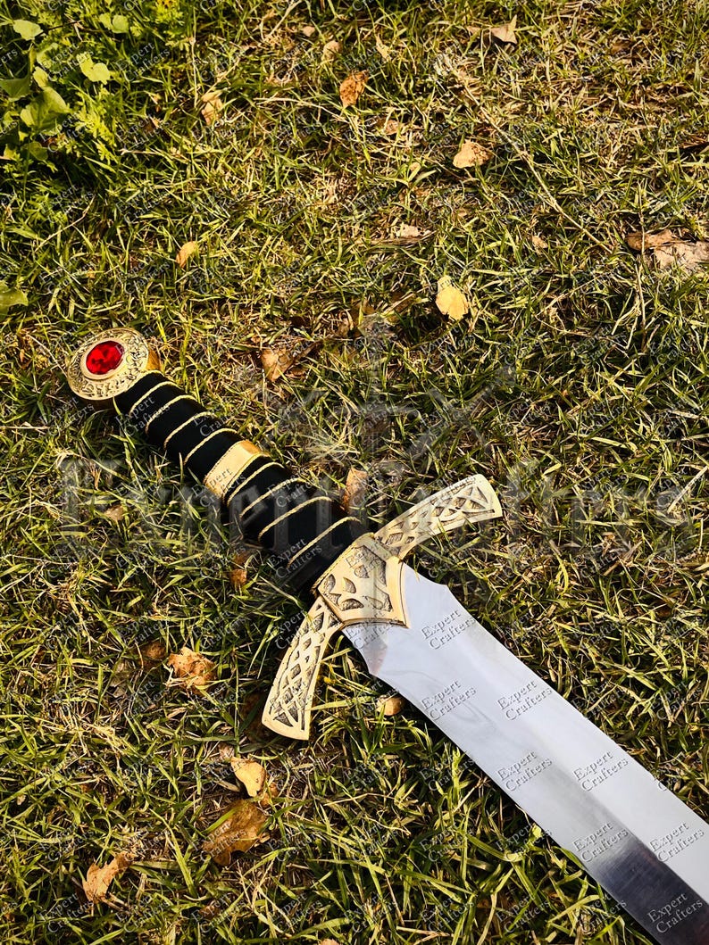 May include: A decorative sword with a silver blade and gold-colored hilt. The hilt features a black handle with gold accents and a red gem at the pommel. The sword is lying on a bed of green grass.