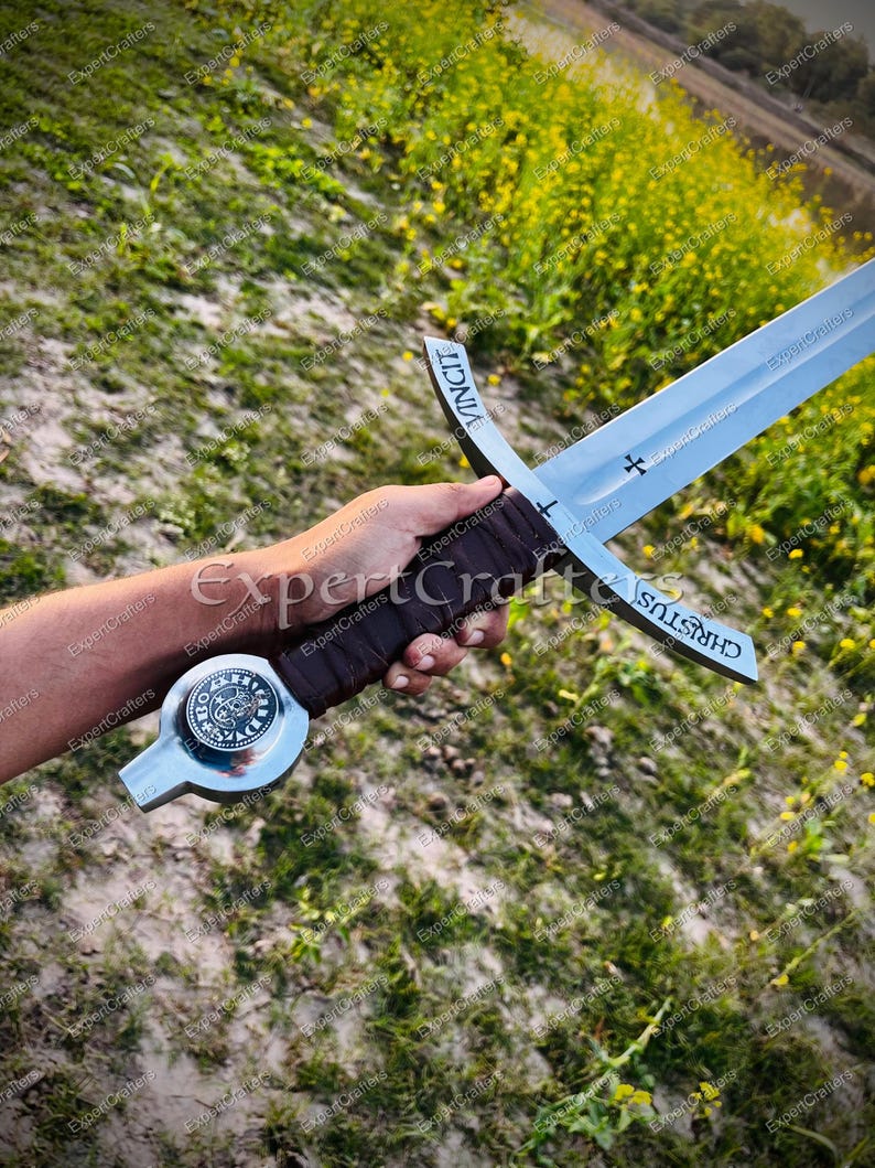 May include: A medieval-style sword with a silver blade and a brown leather-wrapped handle. The crossguard features the word "CHRISTUS" and a cross symbol. The pommel has a circular design. The word "VINCIT" is inscribed on the blade.