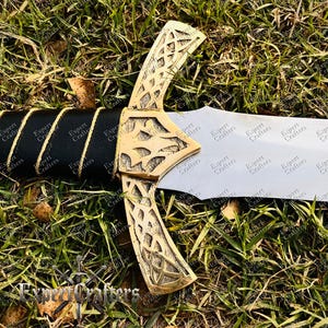 May include: A decorative sword with a gold-colored hilt featuring an intricate Celtic knot design. The handle is wrapped in black leather with gold-colored accents. The blade is silver and the sword is resting on a bed of green grass. The text "Expert Crafters" is visible.