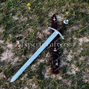 May include: A medieval-style sword with a silver blade and a brown leather-wrapped handle. The sword rests on a bed of grass next to a brown leather sheath adorned with white crosses. The word "CHRISTUS" is etched on the sword.