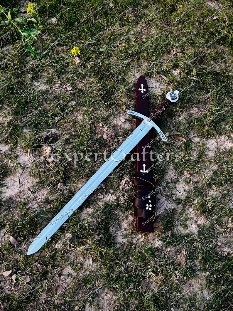May include: A medieval-style sword with a silver blade and a brown leather-wrapped handle. The sword rests on a bed of grass next to a brown leather sheath adorned with white crosses. The word "CHRISTUS" is etched on the sword.