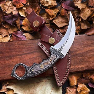 May include: A curved karambit knife with a wooden handle and a leather sheath. The blade is silver with a serrated edge. The handle is gray and brown with brass accents. The sheath is brown with a button closure.