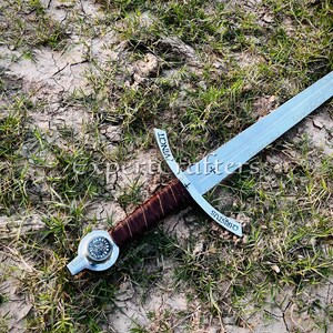 May include: A medieval-style sword with a silver blade and a brown leather-wrapped handle. The crossguard has the word "CHRISTUS" etched on it. The sword rests on a bed of dry grass and dirt.