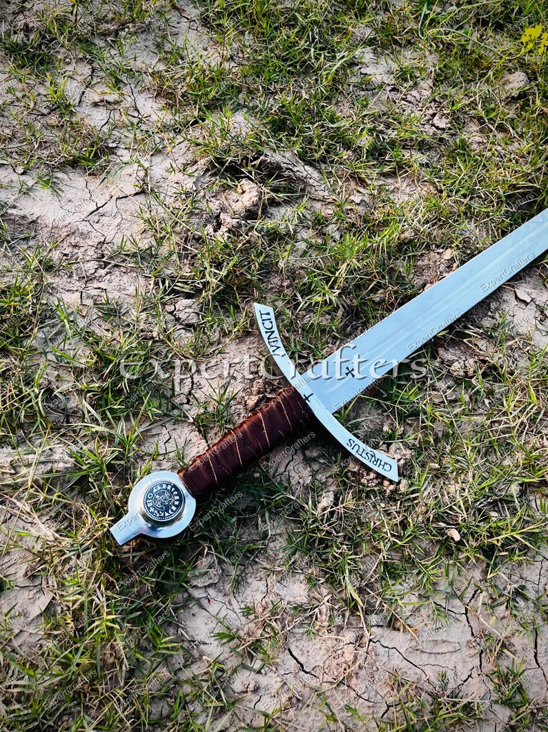 May include: A medieval-style sword with a silver blade and a brown leather-wrapped handle. The crossguard has the word "CHRISTUS" etched on it. The sword rests on a bed of dry grass and dirt.