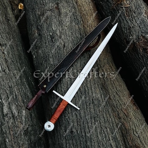May include: A medieval sword with a silver blade and a wooden handle, resting on a dark wooden surface. The sword is accompanied by a black leather sheath. The handle has a crossguard and a decorative pommel.