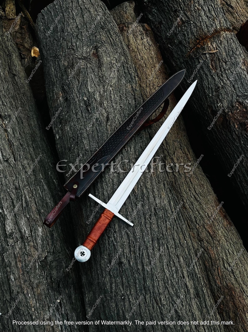 May include: A medieval sword with a silver blade and a wooden handle, resting on a dark wooden surface. The sword is accompanied by a black leather sheath. The handle has a crossguard and a decorative pommel.