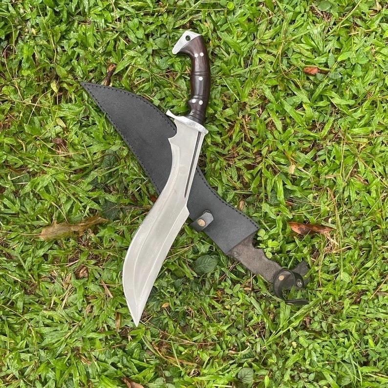 May include: A large, curved blade knife with a dark wooden handle and a silver blade, resting in a black leather sheath. The knife is displayed on a bed of green grass, showcasing its sharp design and craftsmanship.