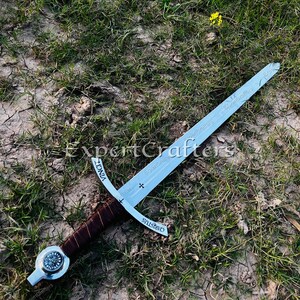 May include: A medieval sword with a silver blade and a brown leather-wrapped handle. The sword features a cross-shaped guard and is inscribed with the words "VINCI" and "CHRISTUS". The sword is lying on a bed of grass and dirt.