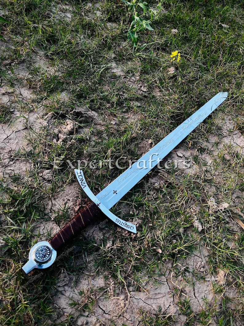 May include: A medieval sword with a silver blade and a brown leather-wrapped handle. The sword features a cross-shaped guard and is inscribed with the words "VINCI" and "CHRISTUS". The sword is lying on a bed of grass and dirt.
