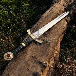 May include: A decorative sword with a silver blade and a black handle wrapped in leather. The hilt is gold-colored with intricate designs, and a red jewel is set in the pommel. The sword rests on a weathered log.