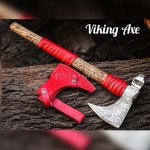 Könnte beinhalten: Eine Wikingeraxt mit Holzgriff und silberner Klinge. Der Griff ist mit rotem Leder umwickelt. Der Axtkopf weist aufwendige Gravuren auf. Eine rote Lederscheide ist ebenfalls vorhanden.