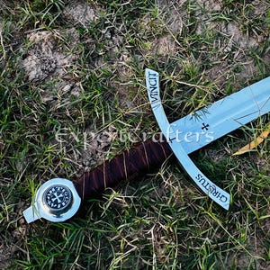 May include: A medieval-style sword with a silver blade and cross-shaped hilt. The hilt is wrapped in brown leather. The sword lies on a bed of green grass. The hilt has the word "VINCIT" and "CHRISTUS" on it.