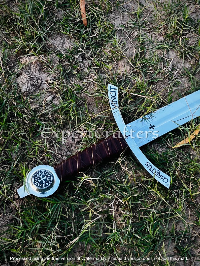 May include: A medieval-style sword with a silver blade and cross-shaped hilt. The hilt is wrapped in brown leather. The sword lies on a bed of green grass. The hilt has the word "VINCIT" and "CHRISTUS" on it.
