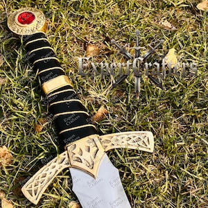 May include: A decorative sword with a gold-colored hilt and crossguard, and a black handle. The pommel features a red gem. The sword rests on a bed of green grass. The text "Expert Crafters" is visible on the sword.