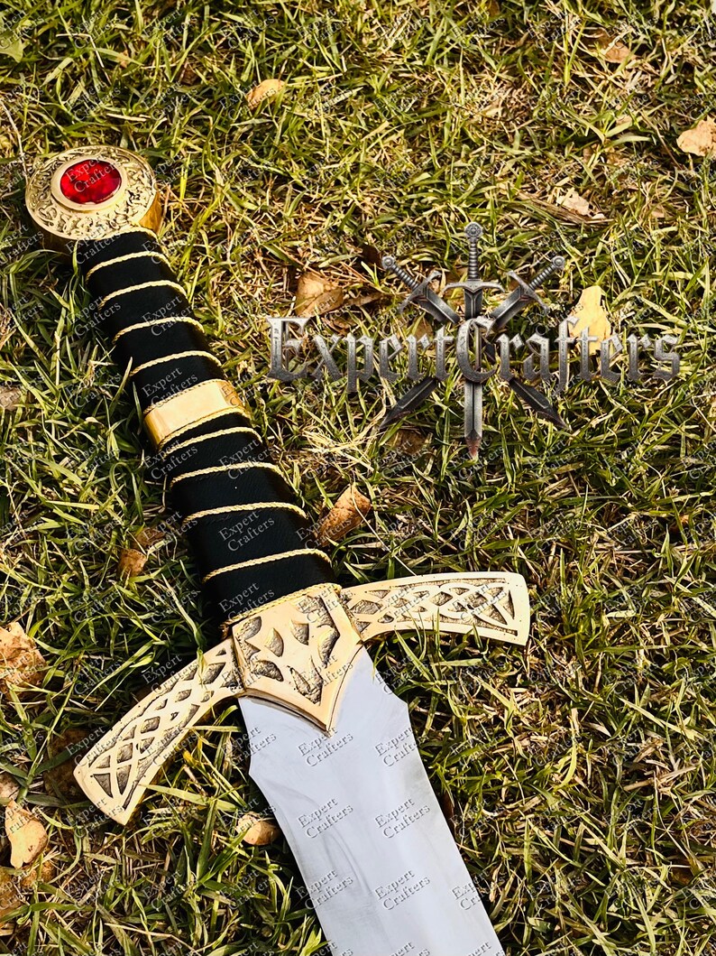 May include: A decorative sword with a gold-colored hilt and crossguard, and a black handle. The pommel features a red gem. The sword rests on a bed of green grass. The text "Expert Crafters" is visible on the sword.