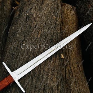 May include: A long, silver sword with a cross-shaped hilt and a brown handle. The blade is sharp and pointed, and the sword is resting on a textured, brown surface. The text "ExpertCrafters" is visible on the blade.