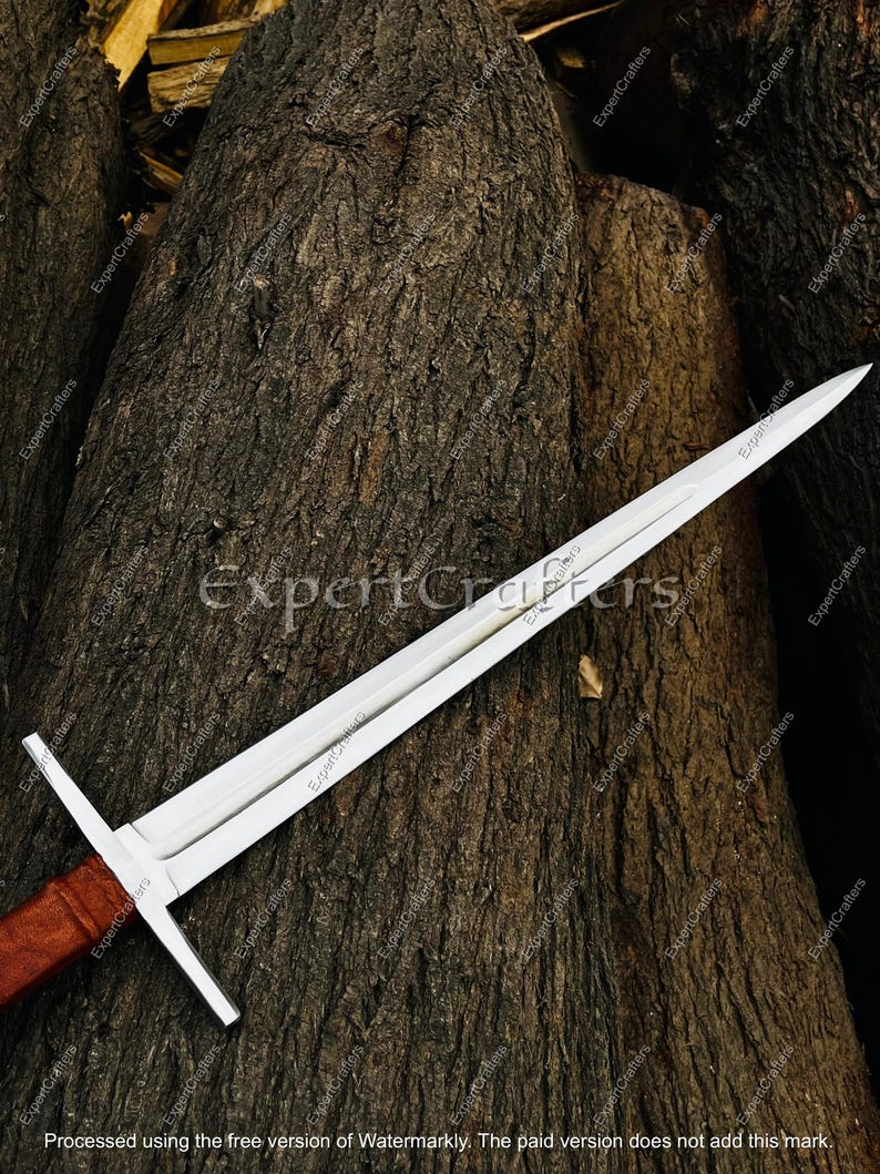 May include: A long, silver sword with a cross-shaped hilt and a brown handle. The blade is sharp and pointed, and the sword is resting on a textured, brown surface. The text "ExpertCrafters" is visible on the blade.