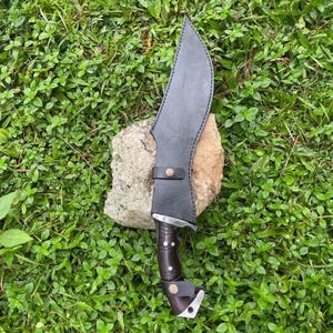 May include: A large, curved knife with a dark wooden handle and a black leather sheath. The blade is silver and the sheath has a strap with a button closure. The knife rests on a rock, surrounded by green foliage.