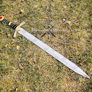 May include: A decorative sword with a silver blade and a gold crossguard. The handle is black and gold, topped with a red gem. The sword lies on a bed of green grass.