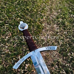 May include: A medieval-style sword with a polished steel blade and crossguard. The handle is wrapped in brown leather. The words "CHRISTUS" and "VINCIT" are etched on the crossguard. The sword is set against a background of green grass.