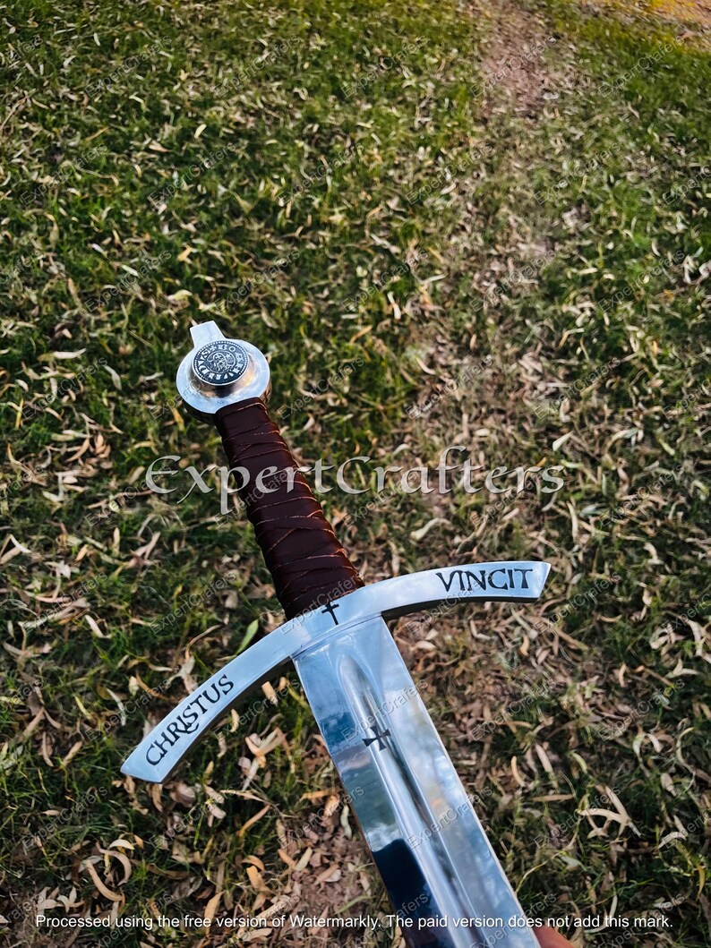 May include: A medieval-style sword with a polished steel blade and crossguard. The handle is wrapped in brown leather. The words "CHRISTUS" and "VINCIT" are etched on the crossguard. The sword is set against a background of green grass.