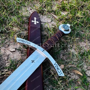 May include: A medieval sword with a silver blade and a brown leather-wrapped handle. The blade has the word "CHRISTUS" and "VINCIT" etched on it, along with cross symbols. The sword rests on a brown sheath with a cross emblem.