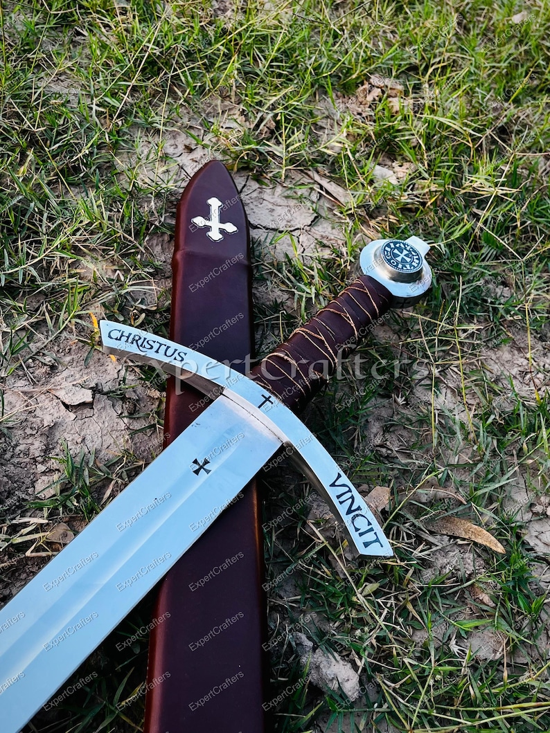 May include: A medieval sword with a silver blade and a brown leather-wrapped handle. The blade has the word "CHRISTUS" and "VINCIT" etched on it, along with cross symbols. The sword rests on a brown sheath with a cross emblem.