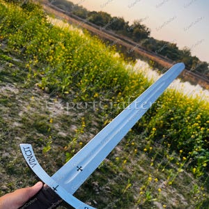 May include: A silver sword with a cross-shaped hilt and a dark brown leather-wrapped handle. The blade has the words "CHRISTUS" and "VINCIT" inscribed on it. The sword is held against a backdrop of a field with yellow flowers and a body of water.