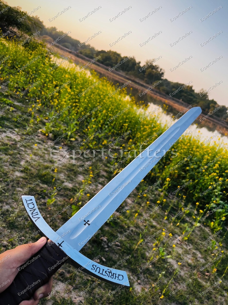 May include: A silver sword with a cross-shaped hilt and a dark brown leather-wrapped handle. The blade has the words "CHRISTUS" and "VINCIT" inscribed on it. The sword is held against a backdrop of a field with yellow flowers and a body of water.
