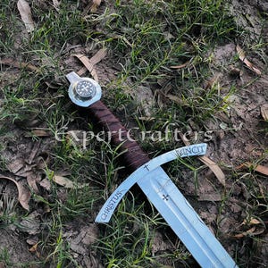 May include: A medieval-style sword with a silver blade and a brown leather-wrapped handle. The crossguard is engraved with the word "VINCIT" and a cross. The blade has the word "CHRISTUS" engraved on it. The sword is lying on the ground.