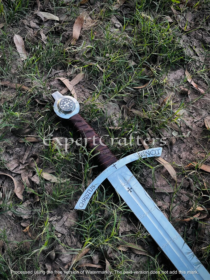May include: A medieval-style sword with a silver blade and a brown leather-wrapped handle. The crossguard is engraved with the word "VINCIT" and a cross. The blade has the word "CHRISTUS" engraved on it. The sword is lying on the ground.