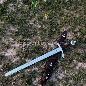 May include: A medieval sword with a silver blade and a brown leather sheath, adorned with white cross symbols. The sword rests on a bed of grass and dirt, with a few yellow flowers in the background.