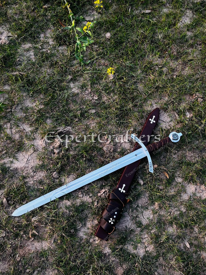 May include: A medieval sword with a silver blade and a brown leather sheath, adorned with white cross symbols. The sword rests on a bed of grass and dirt, with a few yellow flowers in the background.