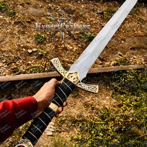 May include: A medieval-style sword with a silver blade and a gold-colored crossguard. The handle is wrapped in black leather with gold accents and a red jewel at the pommel. The text "Expert Crafters" is visible.