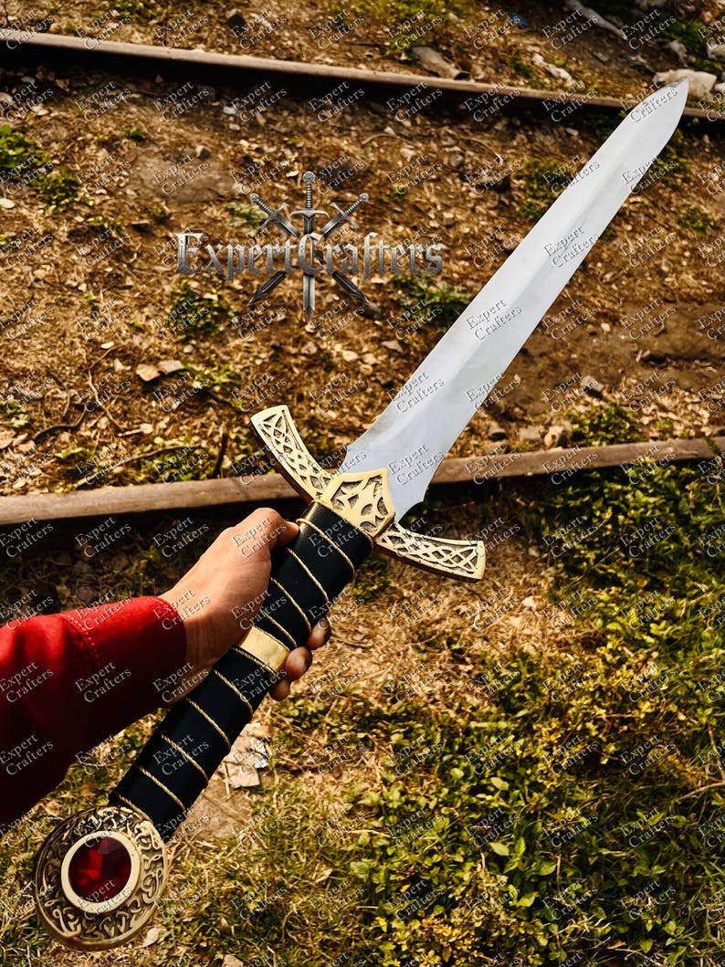 May include: A medieval-style sword with a silver blade and a gold-colored crossguard. The handle is wrapped in black leather with gold accents and a red jewel at the pommel. The text "Expert Crafters" is visible.