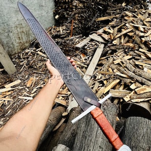 May include: A long, medieval-style sword with a dark, textured blade and a brown leather-wrapped handle. The sword features a cross-shaped hilt and a white pommel with a black cross design. The scabbard is dark brown leather.