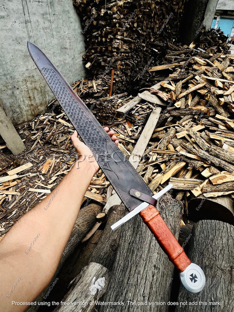 May include: A long, medieval-style sword with a dark, textured blade and a brown leather-wrapped handle. The sword features a cross-shaped hilt and a white pommel with a black cross design. The scabbard is dark brown leather.