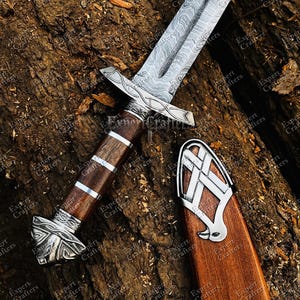 Op de afbeelding: Een Vikingzwaard met een patroon op het lemmet en een houten handvat, versierd met zilverkleurige metalen details. Het zwaard ligt op een getextureerd, bruin oppervlak, met de schede ook zichtbaar.
