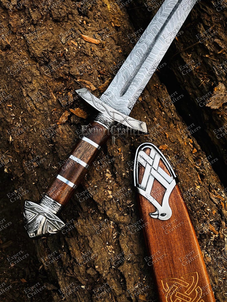 Op de afbeelding: Een Vikingzwaard met een patroon op het lemmet en een houten handvat, versierd met zilverkleurige metalen details. Het zwaard ligt op een getextureerd, bruin oppervlak, met de schede ook zichtbaar.