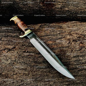 May include: A large, polished hunting knife with a shiny silver blade and a brown leather-wrapped handle. The knife features a brass guard and pommel. The knife is resting on a textured, dark brown wooden surface.
