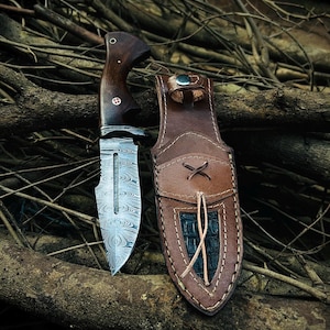 May include: A handcrafted hunting knife with a patterned steel blade and a dark wood handle. The knife is accompanied by a brown leather sheath with a decorative stitched design. The knife and sheath are displayed on a bed of twigs.