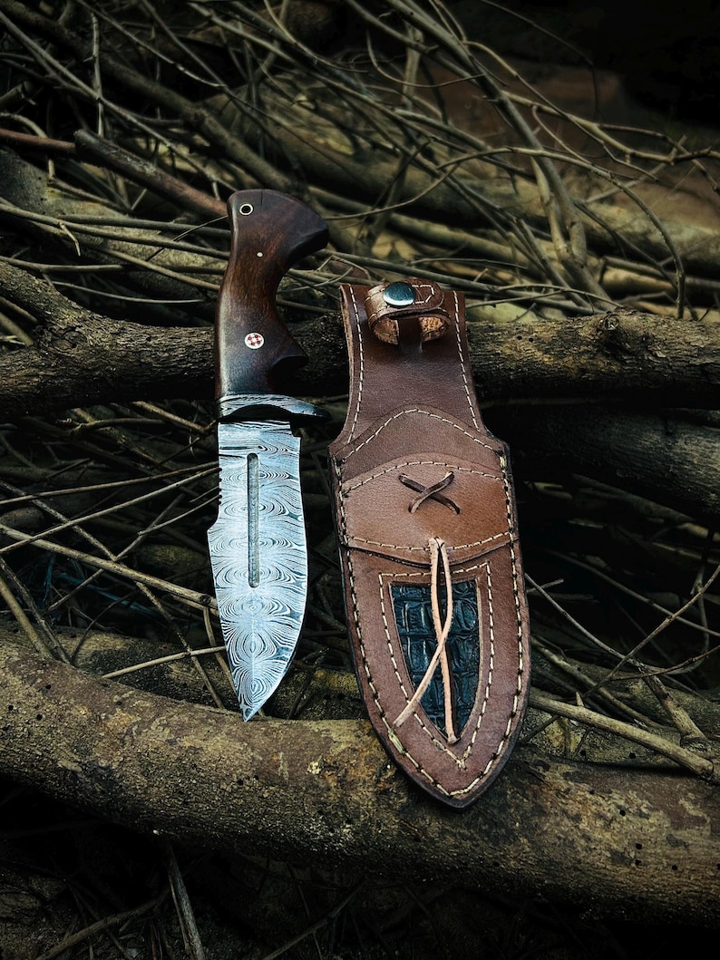 May include: A handcrafted hunting knife with a patterned steel blade and a dark wood handle. The knife is accompanied by a brown leather sheath with a decorative stitched design. The knife and sheath are displayed on a bed of twigs.